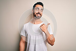 Young handsome man with beard wearing casual t-shirt standing over white background smiling with happy face looking and pointing