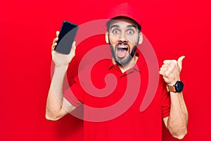 Young handsome man with beard wearing delivery uniform holding smartphone pointing thumb up to the side smiling happy with open