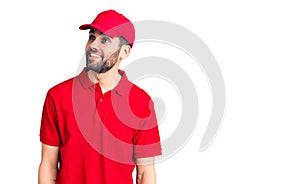 Young handsome man with beard wearing delivery uniform looking away to side with smile on face, natural expression