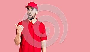 Young handsome man with beard wearing delivery uniform surprised pointing with hand finger to the side, open mouth amazed