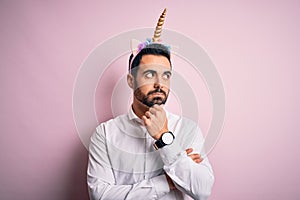 Young handsome man with beard wearing funny unicorn diadem over pink background with hand on chin thinking about question, pensive