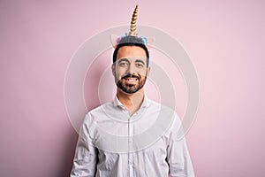 Young handsome man with beard wearing funny unicorn diadem over pink background with a happy and cool smile on face
