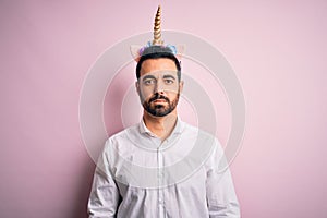 Young handsome man with beard wearing funny unicorn diadem over pink background with serious expression on face