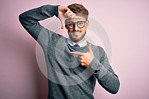 Young handsome man with beard wearing glasses and sweater standing over pink background smiling making frame with hands and