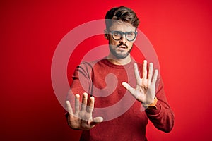 Young handsome man with beard wearing glasses and sweater standing over red background Moving away hands palms showing refusal and