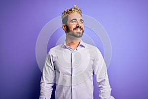 Young handsome man with beard wearing golden crown of king over purple background looking away to side with smile on face, natural