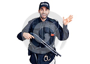 Young handsome man with beard wearing police uniform holding shotgun celebrating victory with happy smile and winner expression