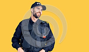 Young handsome man with beard wearing police uniform looking away to side with smile on face, natural expression