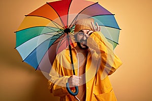 Young handsome man with beard wearing raincoat for rainy day holding colorful umbrella with happy face smiling doing ok sign with