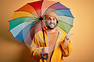 Young handsome man with beard wearing raincoat for rainy day holding colorful umbrella very happy pointing with hand and finger to