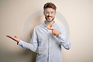 Young handsome man with beard wearing striped shirt standing over white background amazed and smiling to the camera while