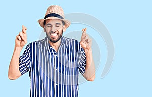 Young handsome man with beard wearing summer hat and striped shirt gesturing finger crossed smiling with hope and eyes closed