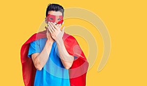 Young handsome man with beard wearing super hero costume shocked covering mouth with hands for mistake