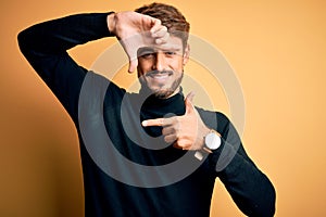 Young handsome man with beard wearing turtleneck sweater standing over yellow background smiling making frame with hands and