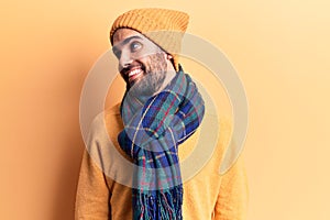 Young handsome man with beard wearing wool cap and scarf looking away to side with smile on face, natural expression