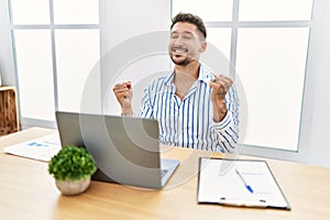 Young handsome man with beard working at the office using computer laptop very happy and excited doing winner gesture with arms
