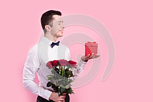 Young handsome man with beautiful flower bouquet and red gift box on pink background