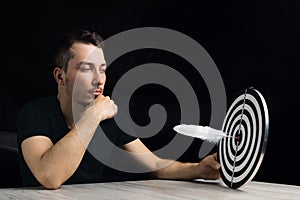 Man sits at table holding dart board with quill pen stuck in the center of the target on black background