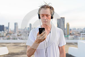 Young handsome man with blond hair using phone while listening to music against view of the city