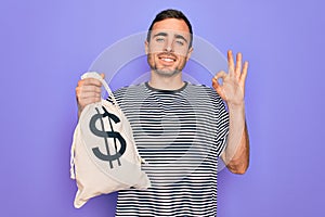 Young handsome man with blue eyes holding bag with dollar symbol over purple background doing ok sign with fingers, excellent