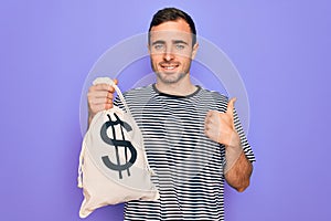 Young handsome man with blue eyes holding bag with dollar symbol over purple background happy with big smile doing ok sign, thumb