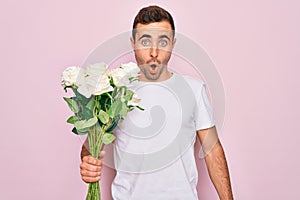 Young handsome man with blue eyes holding bouquet of flowers over pink background scared in shock with a surprise face, afraid and