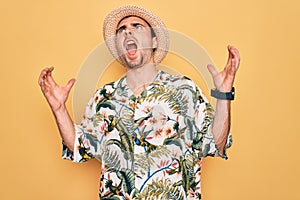 Young handsome man with blue eyes on vacation wearing summer florar shirt and hat crazy and mad shouting and yelling with