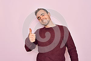 Young handsome man with blue eyes wearing casual sweater standing over pink background doing happy thumbs up gesture with hand