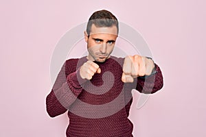 Young handsome man with blue eyes wearing casual sweater standing over pink background Punching fist to fight, aggressive and