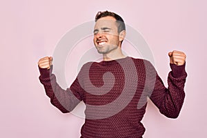 Young handsome man with blue eyes wearing casual sweater standing over pink background very happy and excited doing winner gesture
