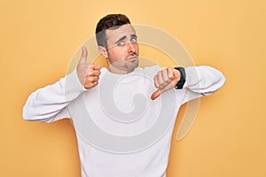 Young handsome man with blue eyes wearing casual sweater standing over yellow background Doing thumbs up and down, disagreement
