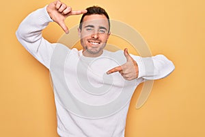Young handsome man with blue eyes wearing casual sweater standing over yellow background smiling making frame with hands and