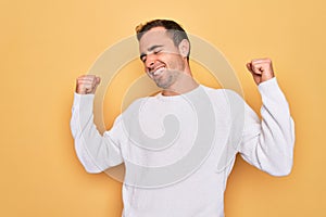 Young handsome man with blue eyes wearing casual sweater standing over yellow background very happy and excited doing winner