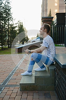 A young handsome man with bristles sits on a granite staircase near a brick building on a summer evening