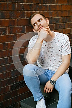 A young handsome man with bristles sits on a granite staircase near a brick building on a summer evening