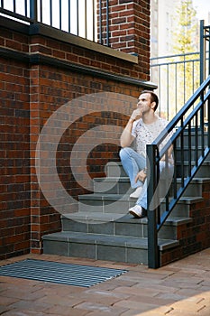 A young handsome man with bristles sits on a granite staircase near a brick building on a summer evening