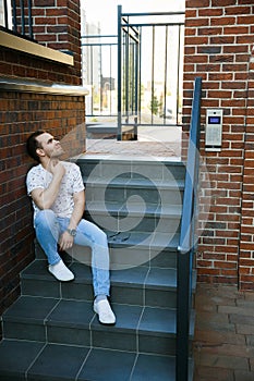 A young handsome man with bristles sits on a granite staircase near a brick building on a summer evening