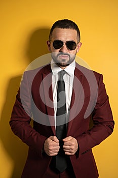 Young handsome man in burgundy suit looking on you seriously wearing sunglasses with hands holding a jacket isolated on