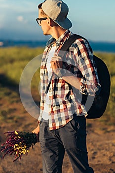 Young handsome man, in a cap and glasses, with a backpack, traveling and walking in nature, a man with a bouquet of wildflowers