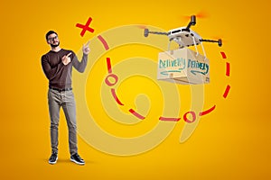 Young handsome man in casual clothes standing and pointing at delivery drone carrying big package, with red routing line