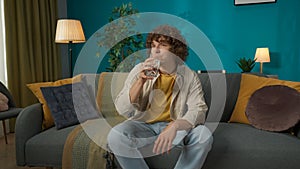 Young handsome man in casual clothing, sitting on the couch in the living room and drinking a glass of water