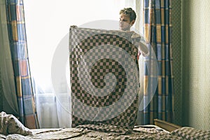 Young handsome man in casual makes the bed holding blanket in the morning against the sunlight throung the window f