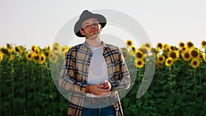 A young handsome man with a cowboy hat is posing in front of his sunflower field and smiling cheerfully as he is looking