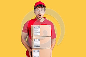 Young handsome man with curly hair holding delivery package scared and amazed with open mouth for surprise, disbelief face