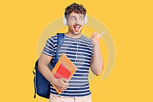 Young handsome man with curly hair holding student backpack and books pointing thumb up to the side smiling happy with open mouth