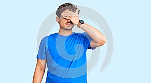 Young handsome man with curly hair wearing casual clothes covering eyes with hand, looking serious and sad