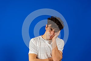 Young handsome man with curly hair wearing white shirt over blue background with hand on face thinking about question