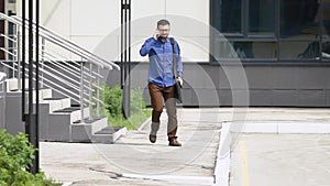 Young handsome man dressed in blue casual shirt and brown trousers is stepping outside from the building speaking on the