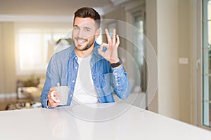 Young handsome man drinking a cup of coffee at home doing ok sign with fingers, excellent symbol