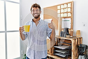 Young handsome man drinking a cup coffee at the office smiling happy and positive, thumb up doing excellent and approval sign